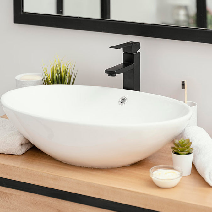 Modern bathroom with white sink on wooden table, black framed mirror, black faucet, potted plant, candle, soap dispenser, white bowl, and towel.
